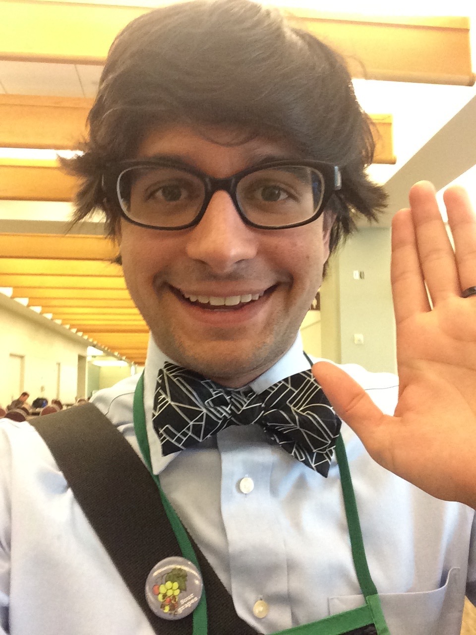 A selfie in a conference center hallway from the chest up of Zhian Kamvar with shaggy dark brown hair, thick black-rimmed oval glasses, a blue button down shirt, and a black bow tie with random white lines traversing it. He is wearing a green conference lanyard and a strap of a shoulder bag with a button on it is also visible. His face shows youthful exuberance as he smiles into the camera and holds up one hand much like agent Dale Cooper would do on the TV series 'Twin Peaks'