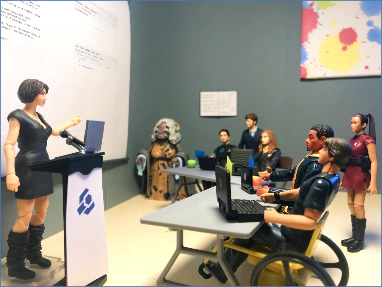 Action figures in a workshop with a lead instructor on the left standing at a podium with The Carpentries logo and a laptop, gesturing to her computer scren projected behind her. There are four learners and two helpers in the audience who all have varying races, abilities, and ages.