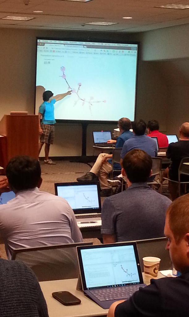 A partially-illuminated workshop classroom from the back of the room. In the foreground are several people sitting at desks with laptops open, all showing the same RStudio window. There are coffee cups on the tables. In the background, Zhian Kamvar is wearing a bright blue polo shirt and checkered shorts as he reaches up and points to a twiggy-looking diagram (minimum spanning tree) displayed on the projector screen.